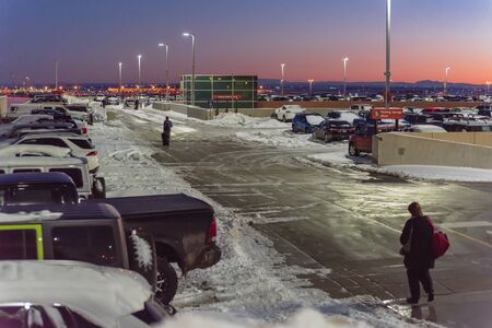 Rear view travelers in warm clothes walking with luggage at terminal parking of Denver International Airport (DIA) in frosty cold autumn sunset. Busy lot with snowdrifts, severe weather conditionの写真素材