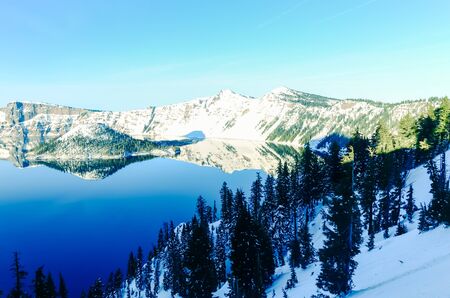 Snow cliff and pine trees lush above Crater Lake with snowcap mountain reflection. During cold snaps fingers of ice probe from the shore out over the abyss. Winter scene at National Park, Oregonの写真素材
