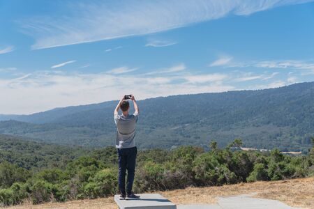 Rear view of young Caucasian man taking photo of nature San Jose mountain landscape from viewpoint, rest area near U.S. 101 Highway. Back of traveler using smart phone take photoの写真素材