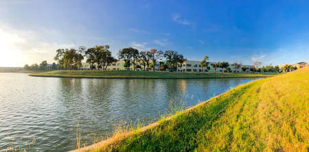 Panorama low angle view from a steep slope hill lawn of lakeside business park in Lewisville, Texas, USA. Waterfront corporate buildings with matured trees under blue cloud sky, early morningの写真素材