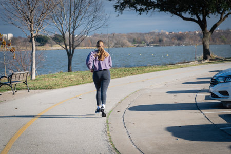 Caucasian lady with earphone running on concrete sidewalk near White Rock Lake in Dallas, Texas,...