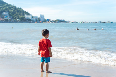 Unidentified 4 year old Asian toddler boy in red t-shirt and short standing on sandy shoreline with Vung Tau City in background. Calm waves and people swimming in sunny morning at tropical oceanの写真素材
