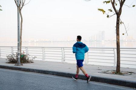 Action motion blurred healthy Asian man with light jacket running along West Lake downtown buildings background early morning in Hanoi, Vietnam. Wellbeing and healthy workout at popular sightseeingの写真素材