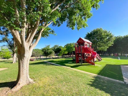 Beautiful suburban park sun poking thru tall matured tree, firetruck themed playground, green artificial grass turf carpet in Carrollton, suburbs Dallas, Texas, USA. Large recreational place for kidsの写真素材