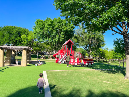 Rear view unidentified kids walking, running at firetruck themed playground with green artificial grass turf carpet, tall trees clear blue sky in Carrollton, suburbs Dallas, Texas, USA. Suburban parkの写真素材