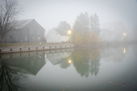 Foggy morning scenic Erie canal with reflection of historic two-story houses, pole lighting and colorful fall foliage riverside walk in Fairport Village, Upstate New York, USA. Scenic small-town Americaの写真素材
