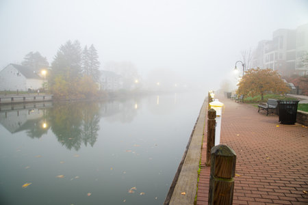 Brick walkway in foggy morning mist along historic Erie Canal with traditional two-story houses, modern apartment complex in both banks in Fairport Village, Upstate New York, USA. Scenic small-townの写真素材
