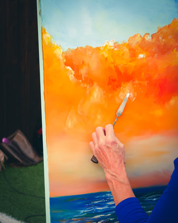 Toned photo of sunset ocean sky painted by senior lady artist hand holding palette knife finishing colorful abstract on easel canvas stand, applying oil acrylic color mixing spreading. Senior painterの写真素材