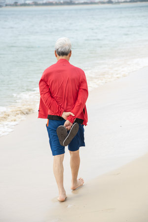 Rear view senior Korean man in red light jacket, short walking barefoot along white sandy beach...