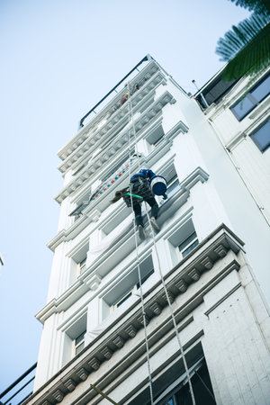 Upward view Asian industrial climber worker at height painting outside high rise building tower in...