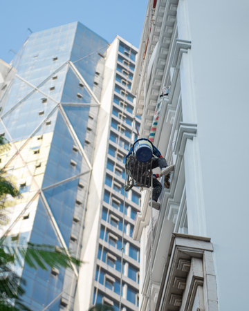 Asian industrial climber worker at height cleaning outside high rise building tower with trees in Hanoi, mountaineering deploys rope access perform tasks at difficult-to-access locations. Vietnamの写真素材