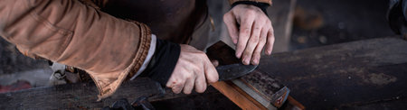 Panorama view blacksmith in leather apron at trade workshop, Mansfield, Missouri, Caucasian craftsman sharpens knife blade on whetstone, sharpening stone, grind edges of steel tool on iron billet. USAの写真素材