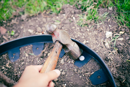 Using hammer to nail galvanized spike pierce through no-dig recycled black plastic landscape edging roll to form tight circle around ring, retaining bedding materials guard fruit tree. USAの写真素材