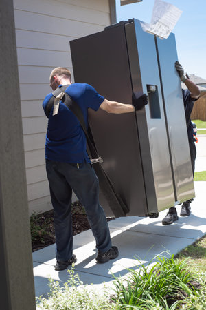 Two workers using shoulder dolly lifting straps, gloves delivering new refrigerator in modern home via concrete walkway in Princeton, Texas. Appliance delivery service truck parked upfront street. USAの写真素材