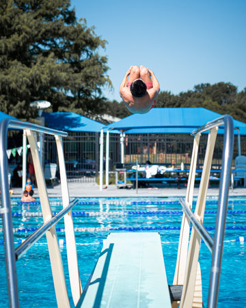 Boy in red swim trunks dives into sunlit outdoor pool beneath bright blue canopies. Captured mid-air with perfect form, image radiates summer energy, motion, and youthful athleticism, Dallas. Texasの写真素材