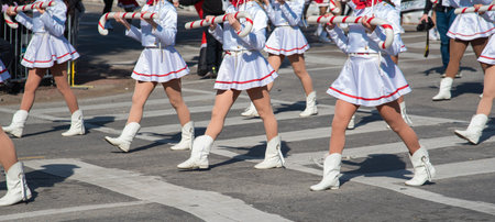 Panorama view parade performers in white skirts, red scarves march oversized J shaped candy cane...
