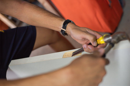 Seated in relaxed posture, craftsman carefully guides yellow-handled chisel along the edge of door wood surface, using hammer to shape mortise hinge with controlled strikes steady hand placement. USAの写真素材