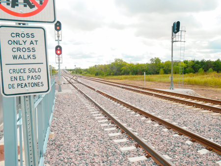 Bilingual sign at rail crossing near DFW Airport urging pedestrians to cross only at designated walkways. Red signal lights and metal railing enhance safety near active tracks, sunny cloud sky. USAの写真素材