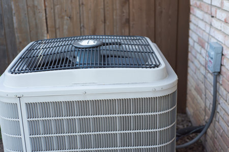 Angled view of AC condenser in Coppell, Texas showing fan grille, electrical conduit, and wall-mounted emergency shutoff switch. Mulch and wooden fence surround the mid-life conditioning unit. USAの写真素材