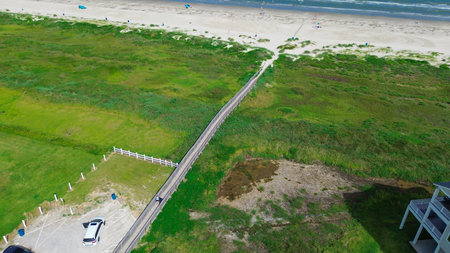 Outdoor parking near elevated beach houses in varied pastel tones line a paved road in Galveston, Texas, grassy dunes and boardwalks separating homes from shoreline and visible beach activity.の写真素材