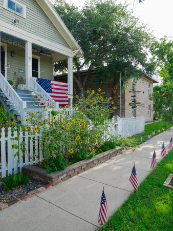 Clean sidewalk lined with American flags runs beside sunflower patch and white picket fence, matured trees, morning lights adds depth to peaceful, patriotic Galveston residential streetscape, TX. USAの写真素材