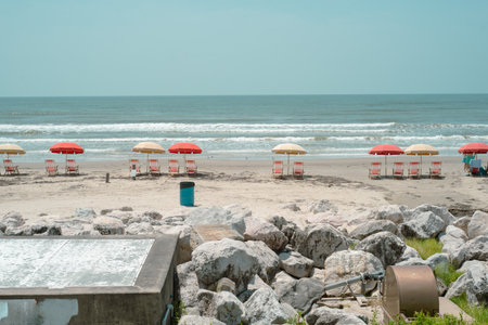 Round metal fixture and exposed pipeline emerge near large white rocks in foreground of quiet Galveston beach, adding industrial texture to scene of neatly arranged umbrellas and calm waves, TX. USAの写真素材