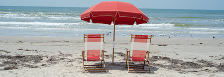 Panorama view calm waves with seagulls by shoreline, red umbrellas striped wooden chairs highlight nostalgic beach aesthetics, focus on intimate seating arrangements, sandy views, Galveston, TX. USAの写真素材