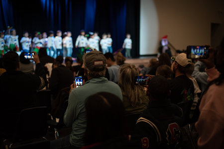 Multiple phones from seated adults attentively record and watch kids performance. The scene reflects...