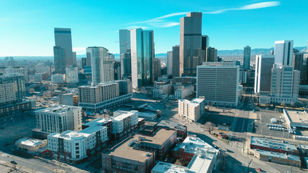 Dense urban cores emerge behind local industrial sites nestled near bustling downtown periphery. Glinting metal surfaces reflect intense solar rays against chilly winter vistas in Denver, CO. USA