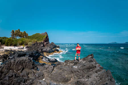 A man in a red shirt stands on a rock by the sea and looks into the distance