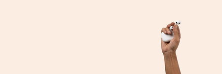 Male hand holding wireless headphones isolated on light pink banner background.