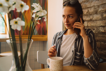 Young pretty woman sitting in a cafe and talking on the phone. The girl drinks hot coffeeの写真素材