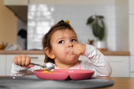 baby toddler girl eats her lunch on the kitchen with spoon. Messy handsの写真素材