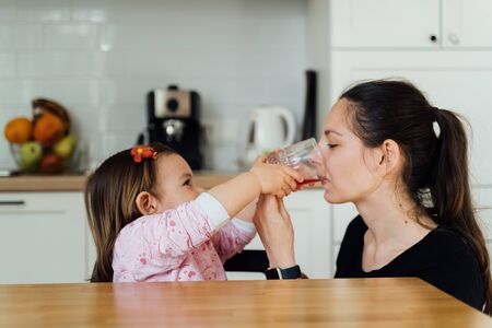 Young woman with baby in kitchen.Drinking a glass of juice. Child sharing beverage with motherの写真素材