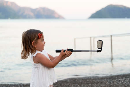 Caucasian small girl with selfie stick on the seaside. Taking photo, recording vlog, video call