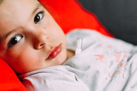 Toddler girl with mercury thermometer lying at home. Kid having high body temperature. Sick, fewer,...