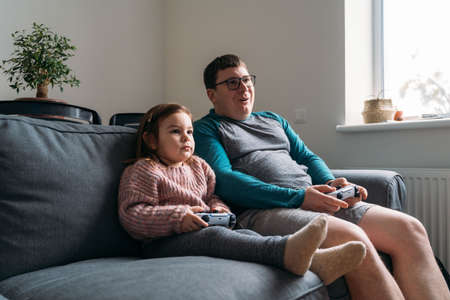 Happy father with child playing wireless console game station. Funny weekend family time at home in...