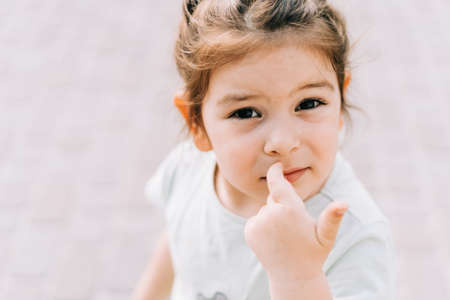 Toddler girl portrait outdoors. Lifestyle child face with big brown eyes. Picking her nose bad habit. Copy space of childhood banner. concerned, serious expressionの写真素材
