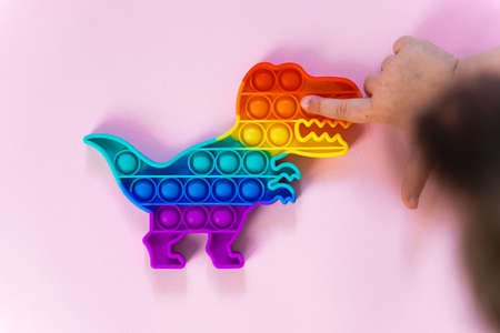 Popit rainbow toy in rainbow colours and dino shape. Trendy bubble pop fidget. Funny anti stress sensory trend. pink background and finger popping the bubbles.の写真素材