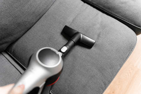 Cleaning grey sofa with wireless vacuum cleaner. Handheld cordless cleaner. Household appliance....