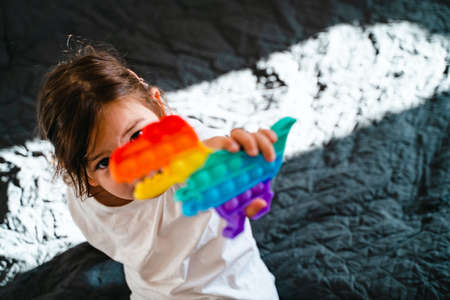 Toddler girl with  toy in rainbow colours. Dino shape silicone toy for stress relief. Bubbles sensory trendy fidget. Child playing at home in candid styleの写真素材