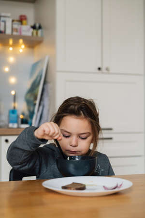 Beautiful child girl eats soup from black bowl with bread and onion. Lifestyle photo of kid in kitchen having a meal.の写真素材