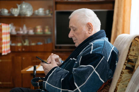 Elderly man with smartphone sitting in an armchair and scrolling social media.