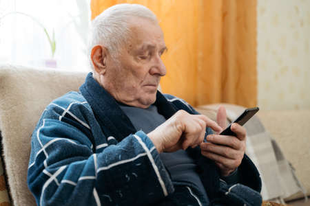 Elderly man with smartphone sitting in an armchair and scrolling social media.
