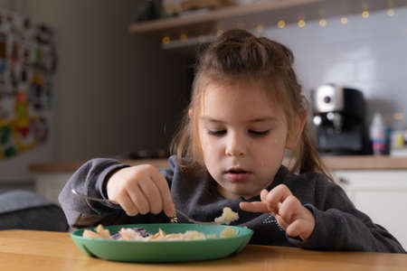 Little child girl sitting at the table and putting spoon at her mouth while eating porridge attentively and with appetite. Stock photoの写真素材