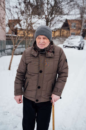 Senior old man caucasian pensioner grandfather holding his cane on the white snow and standing in winter day while looking away with smileの写真素材