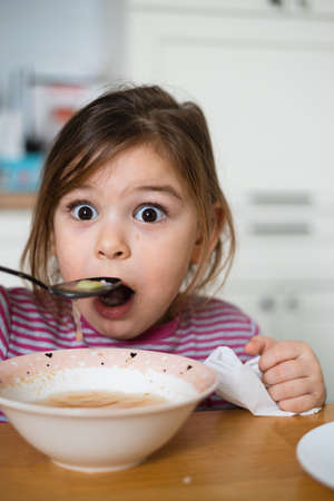 Close up of preschool girl portrait with funny expression eating soup at homeの写真素材