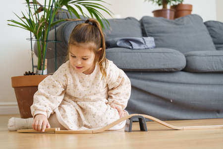 Toddler girl in white dress plays with wooden train at home in the living roomの写真素材