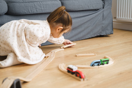 Toddler girl in white dress plays with wooden train at home in the living roomの写真素材