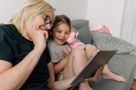 Granny and preschool girl using tablet together at home on the sofa, family togetherness time